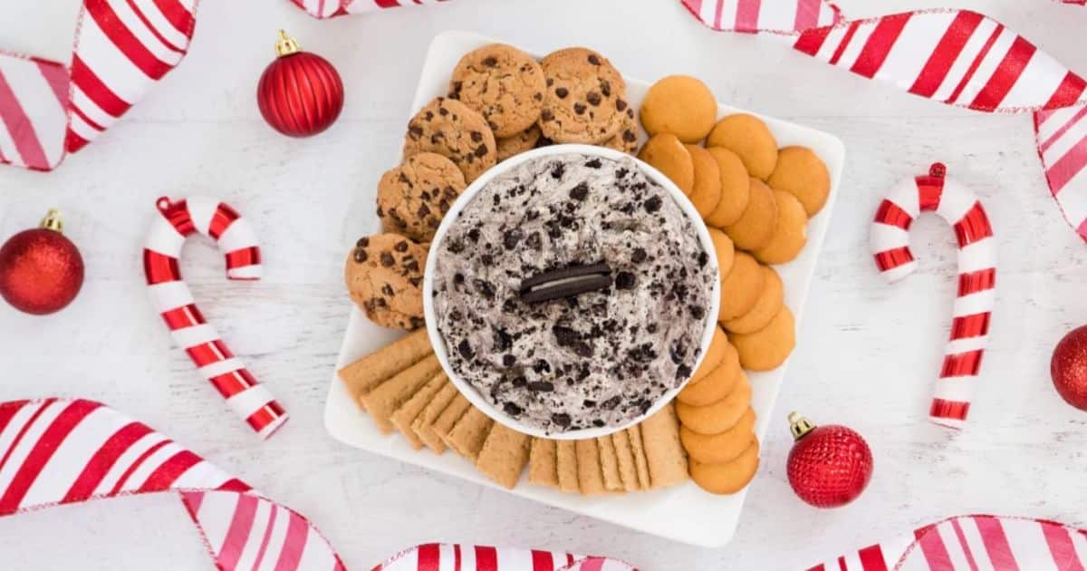 Square platter with cookies and cream dip, assorted cookies, candy canes, and red ornaments on a festive table.