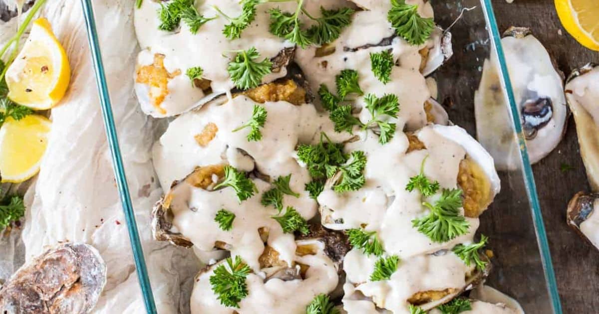 Baked oysters in a glass dish, topped with creamy sauce and fresh parsley, with lemon wedges nearby.