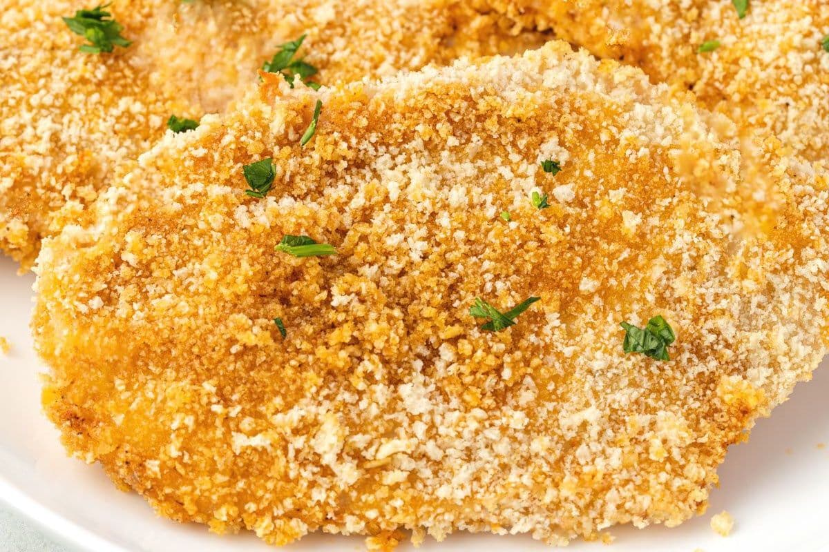 A close-up of crispy, breaded chicken cutlets garnished with chopped parsley on a white plate.