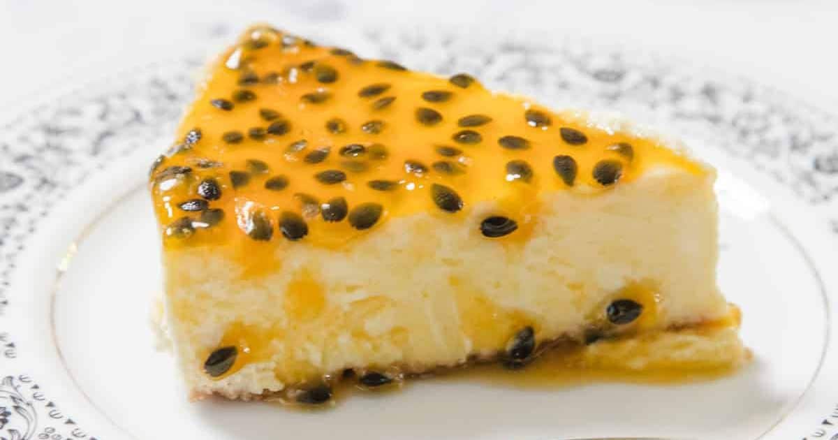 A slice of cheesecake topped with passion fruit sauce and seeds on a white patterned plate.
