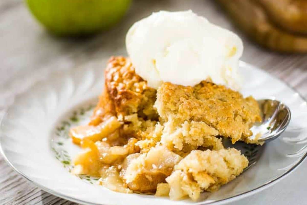 A serving of apple crumble topped with a scoop of vanilla ice cream on a white plate with a spoon.