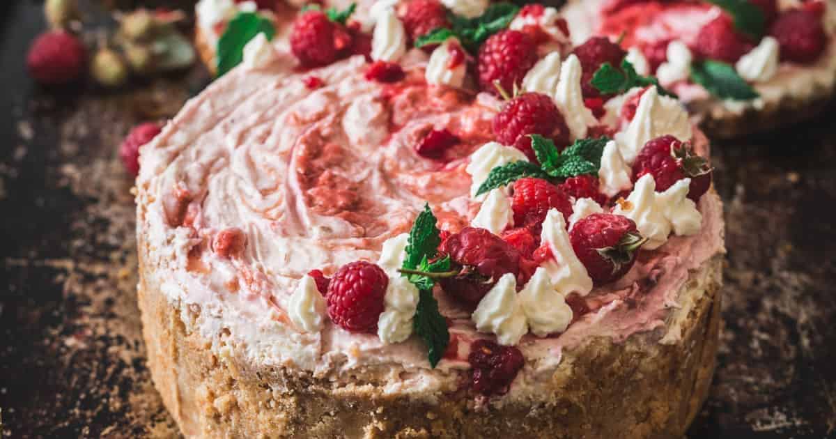 A raspberry cheesecake topped with whipped cream, fresh raspberries, and mint leaves on a dark surface.