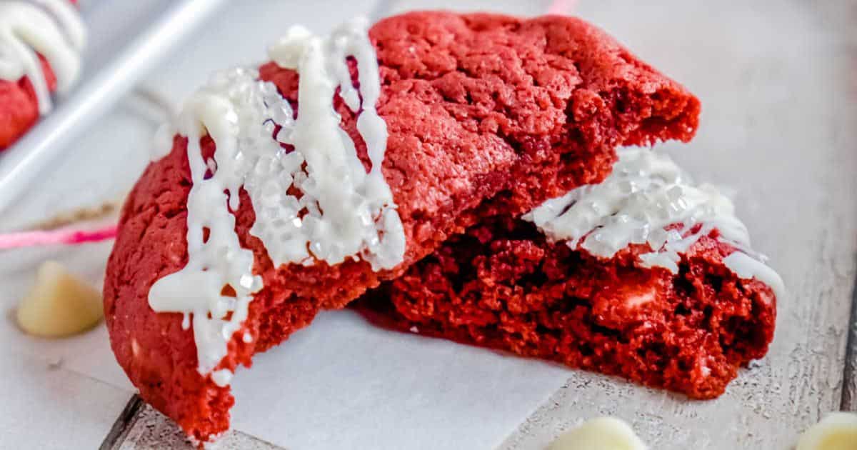 Red velvet cookies with white chocolate drizzle, one broken in half, on a white surface.