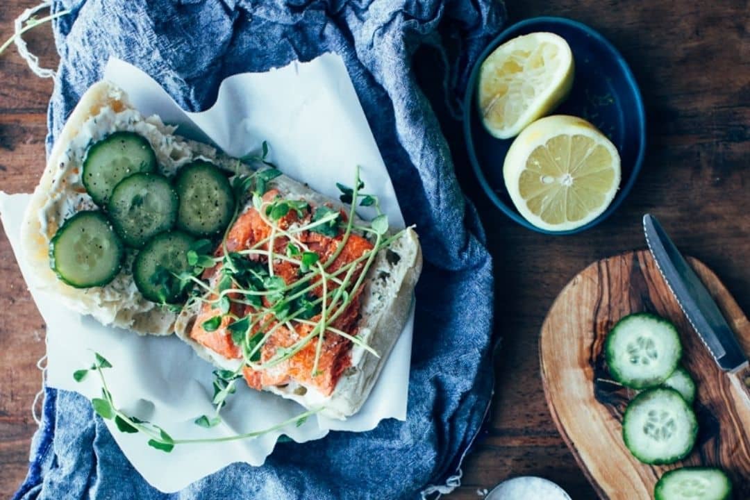 Open sandwich with cucumbers, salmon, microgreens on bread, lemon halves and cucumber slices on the side.