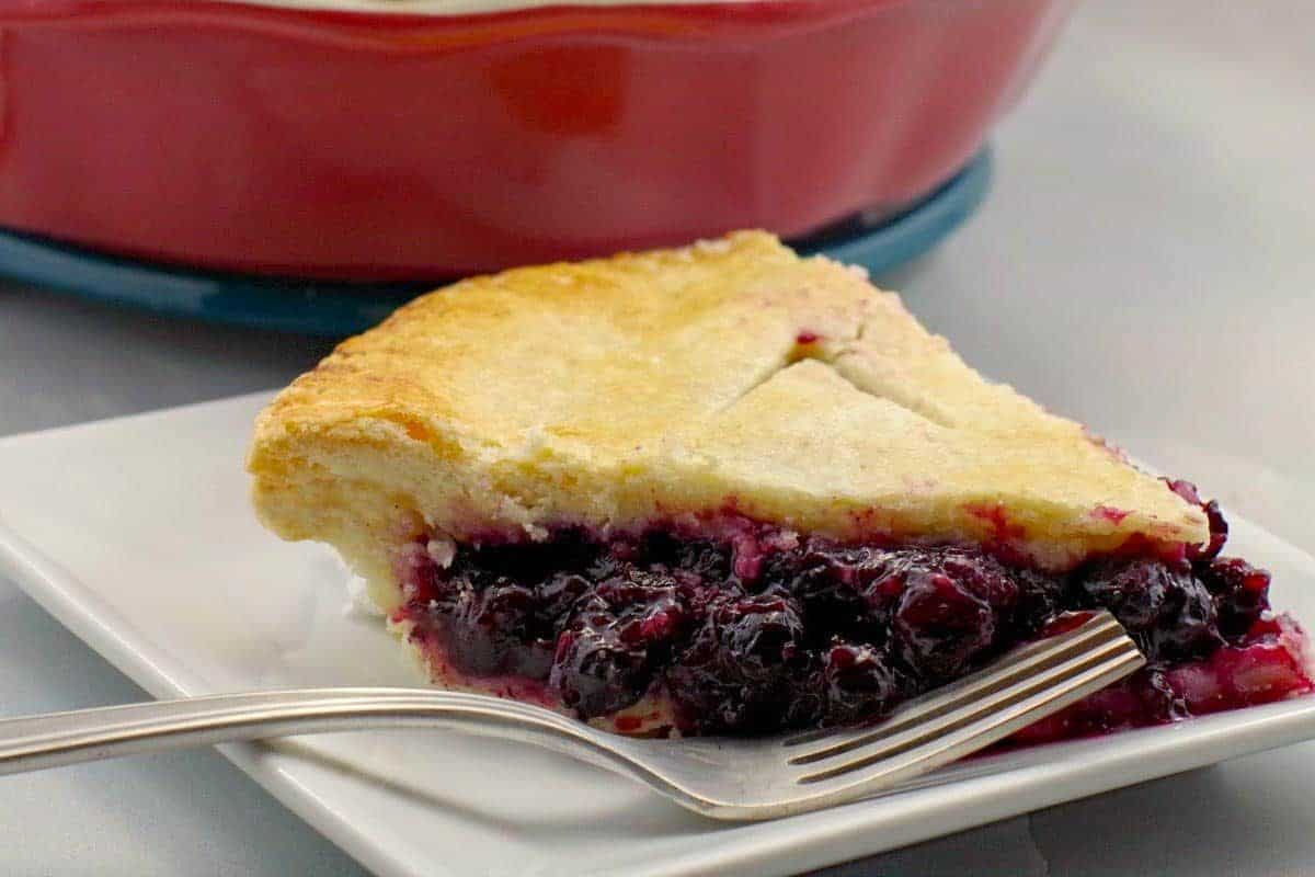 A slice of berry pie with golden crust and a fork on a white plate, with a red dish in the background.
