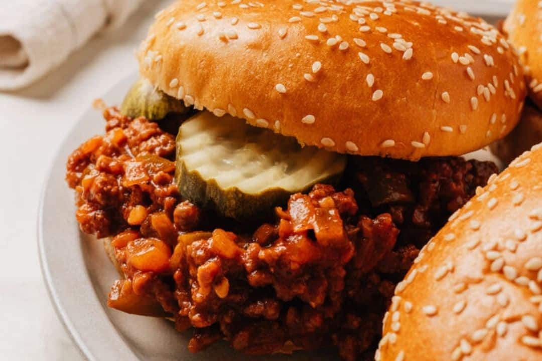 A sloppy joe sandwich with pickles on a sesame seed bun sits on a plate.