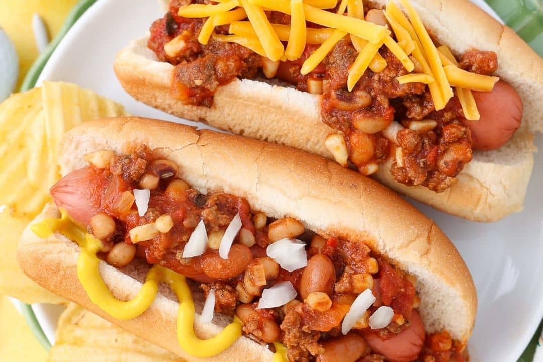 Two chili dogs in buns, one topped with cheese, with chips on the side.