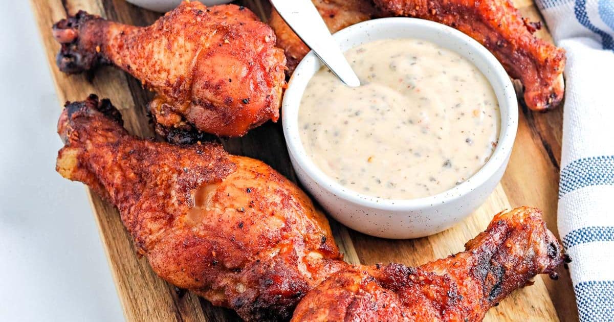 Four seasoned chicken drumsticks on a wooden board with a bowl of creamy dipping sauce and a spoon.