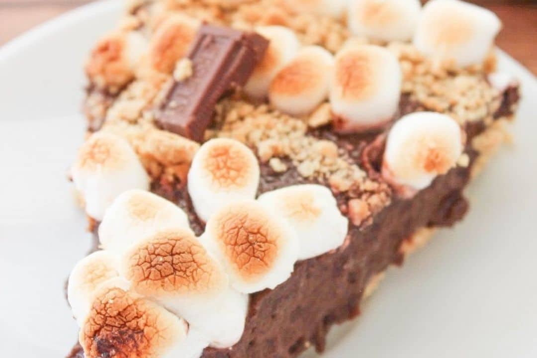 A slice of chocolate pie topped with toasted marshmallows and a piece of chocolate on a white plate.