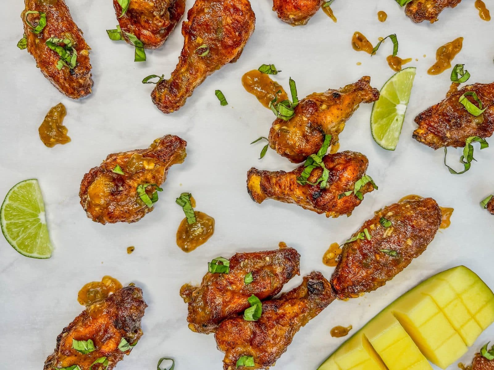Chicken wings garnished with herbs, served with lime wedges and sliced mango on a white surface.