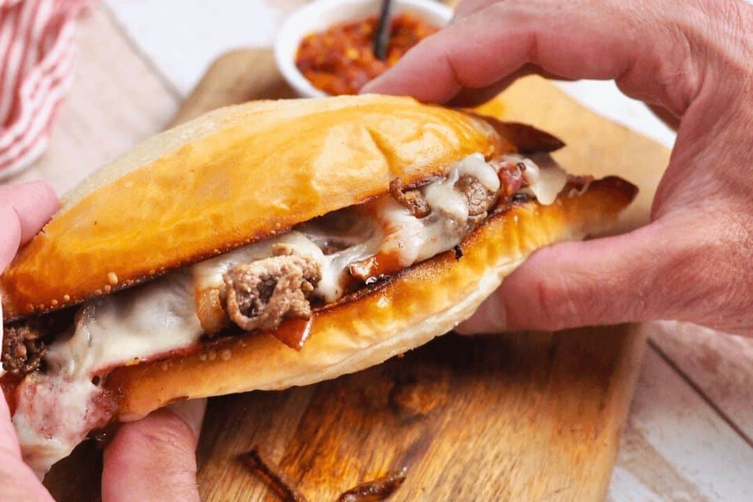 Hands holding a toasted sandwich filled with melted cheese and meat on a wooden board.