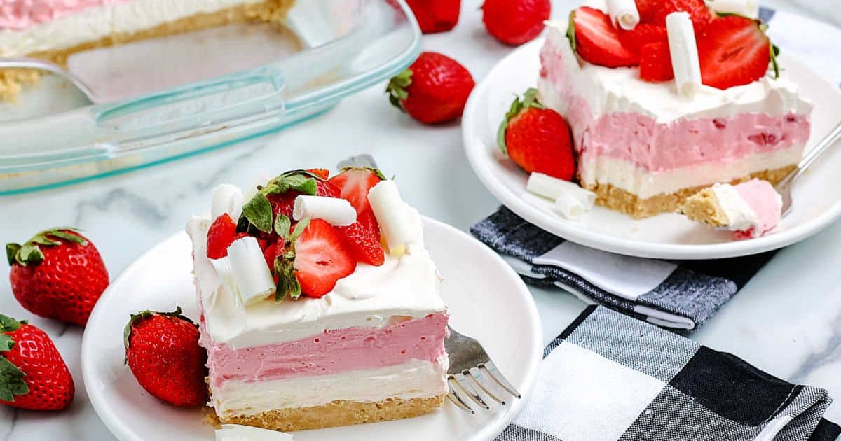 Two plates with layered strawberry dessert slices, topped with whipped cream and fresh strawberries, beside whole strawberries.