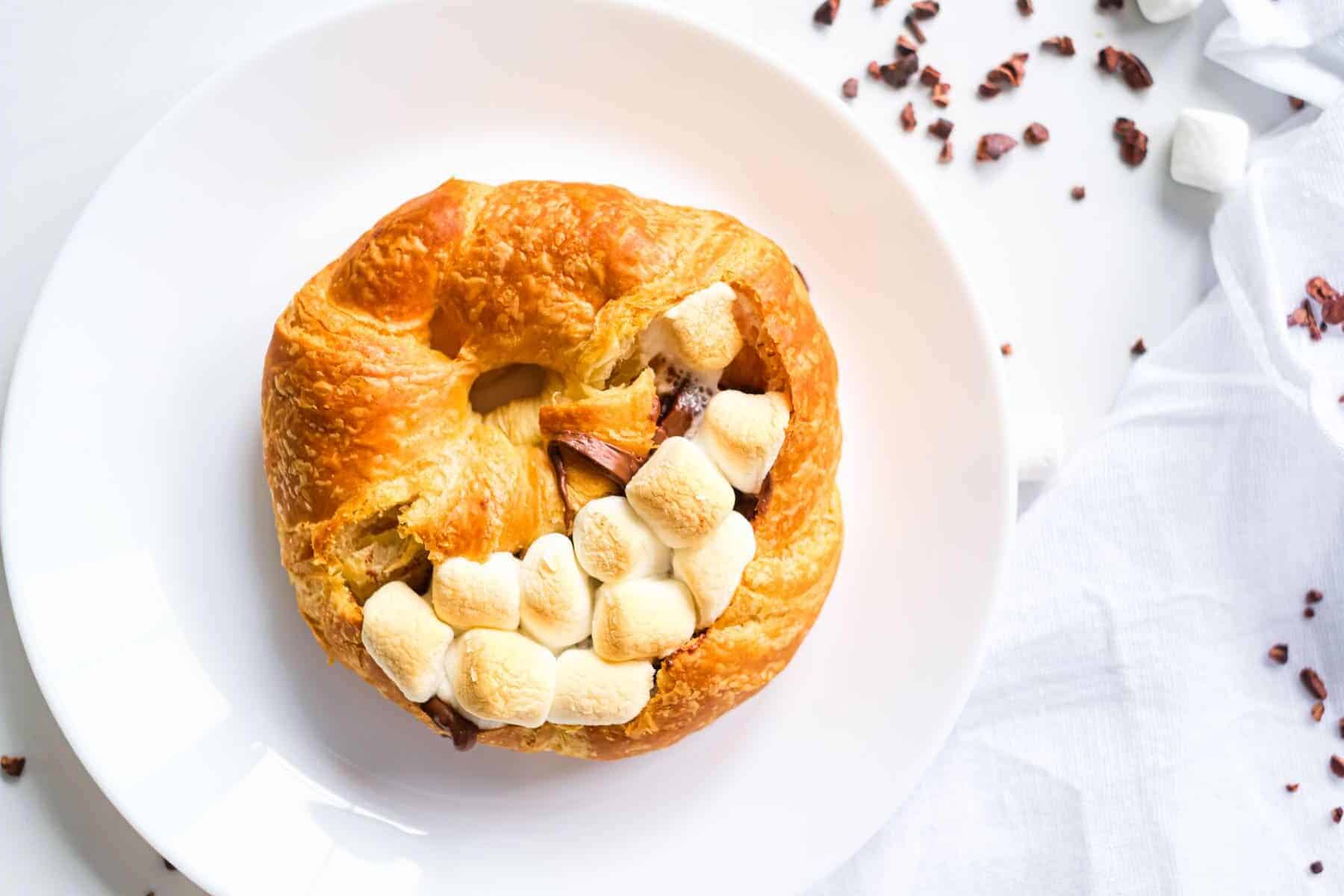 A croissant filled with chocolate and toasted marshmallows on a white plate, with marshmallows scattered nearby.