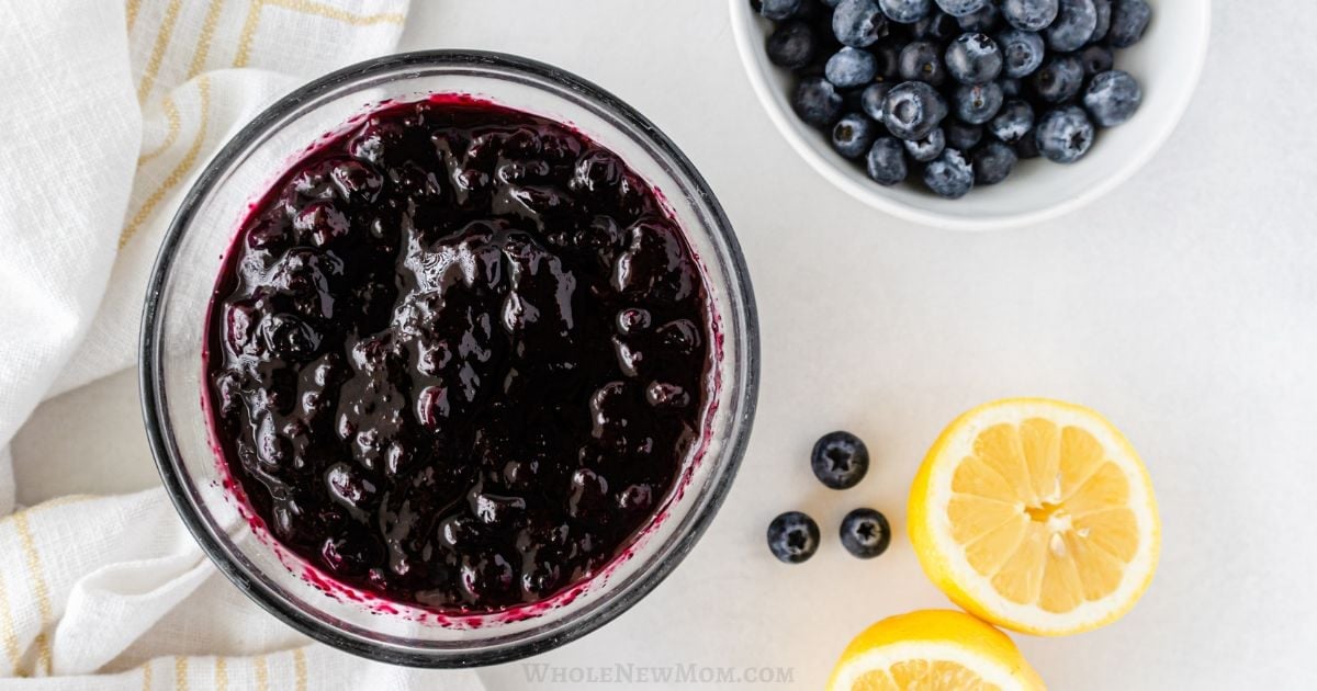 A bowl of blueberry jam next to fresh blueberries and lemon slices on a white surface.