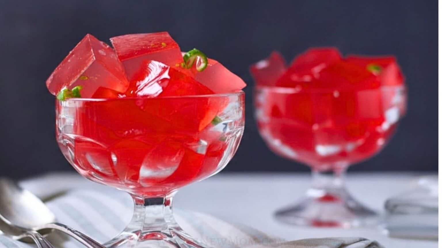 Two glass bowls filled with bright red jelly cubes, placed on a white surface with spoons nearby.
