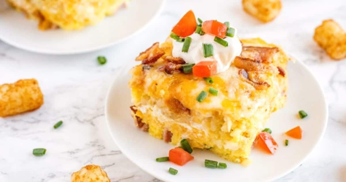 A slice of cheesy breakfast casserole topped with sour cream, chopped tomatoes, and chives on a white plate.