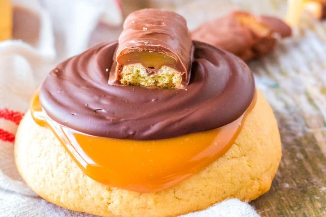 A cookie topped with caramel, chocolate, and a halved chocolate bar with caramel filling.