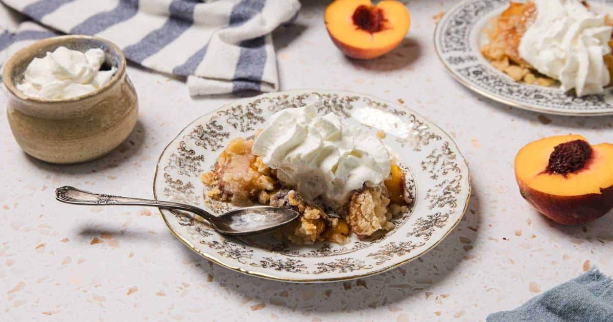 A plate of peach cobbler topped with whipped cream, with a spoon, fresh peach, and whipped cream bowl nearby.
