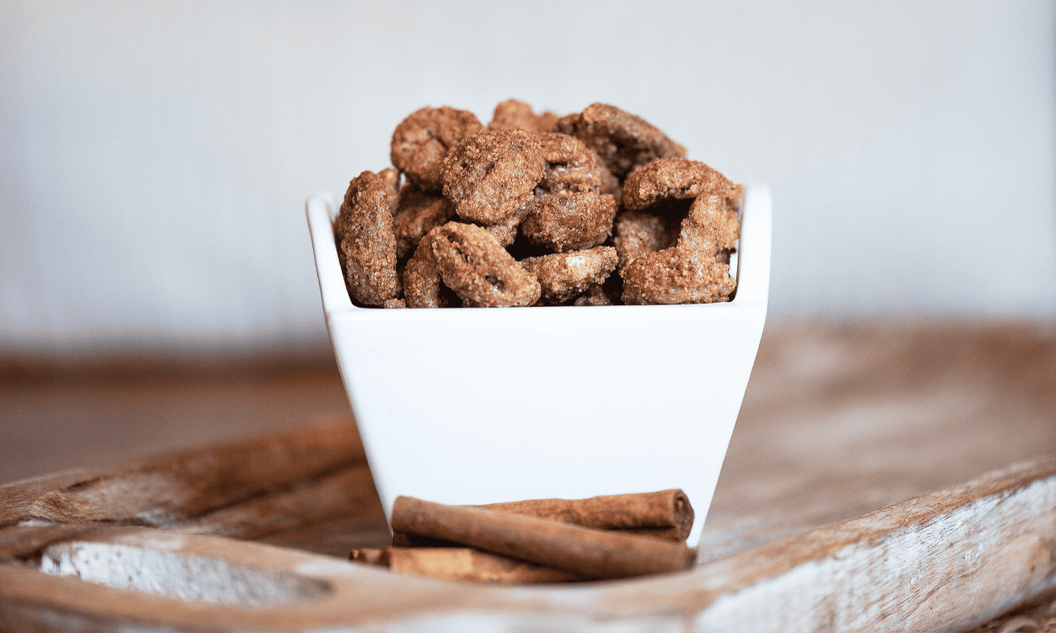A white bowl filled with candied pecans, with cinnamon sticks placed in front on a wooden tray.