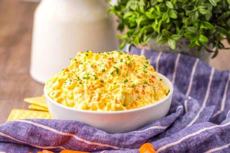 A bowl of egg salad garnished with herbs sits on a blue striped cloth with crackers nearby.