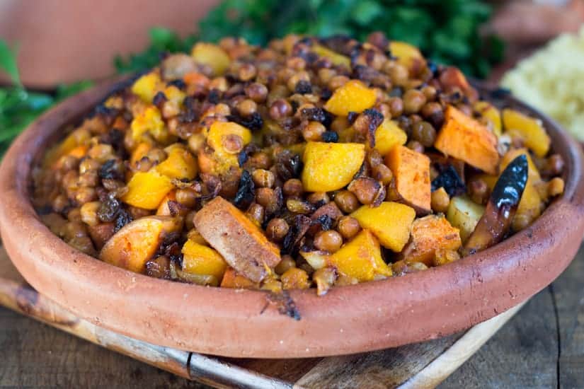 A clay dish filled with cooked chickpeas, vegetables, and spices, served on a wooden board.