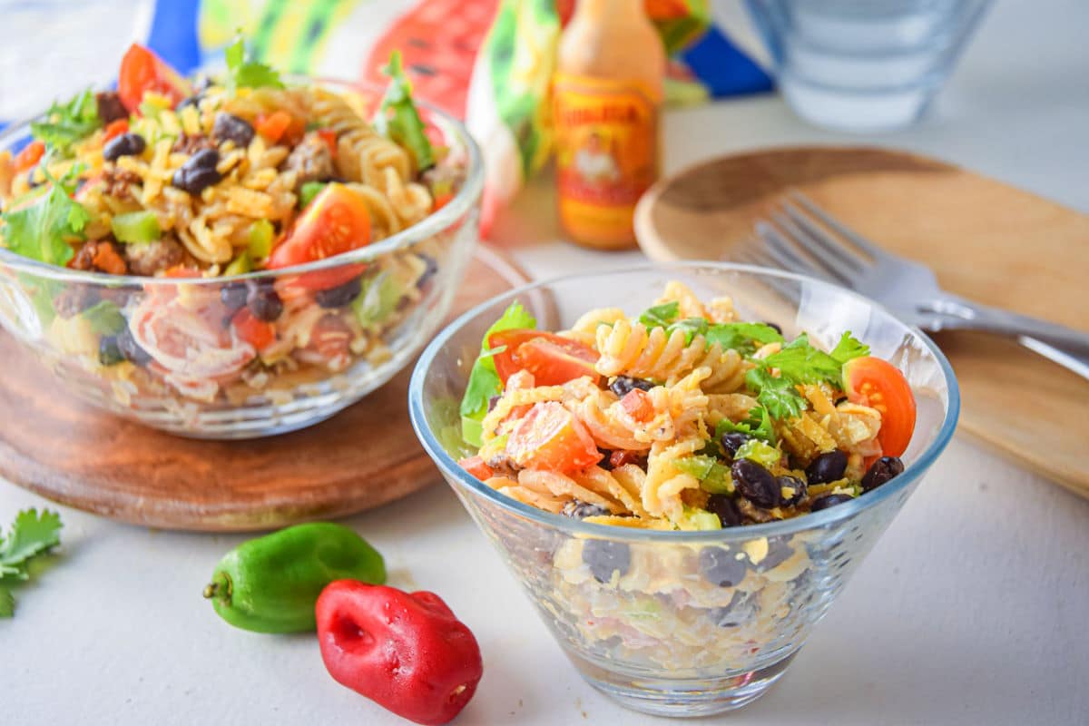 Pasta salad with black beans, tomatoes, and greens in glass bowls, with hot sauce and peppers nearby.