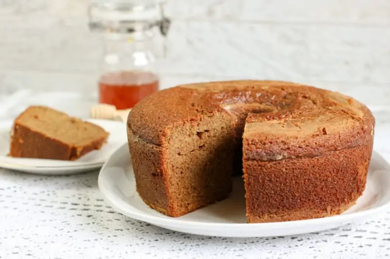 Round honey cake with a slice cut out, served on a white plate, jar of honey in the background.