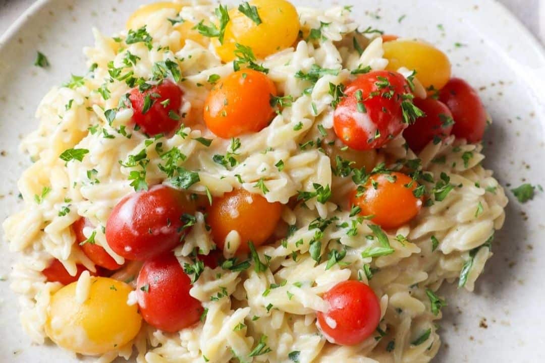 Creamy orzo pasta mixed with yellow and red cherry tomatoes, garnished with chopped parsley on a white plate.