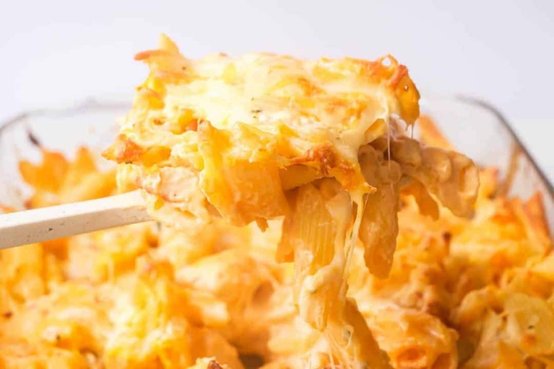A cheesy pasta bake being scooped out of a glass baking dish with melted cheese stretching.
