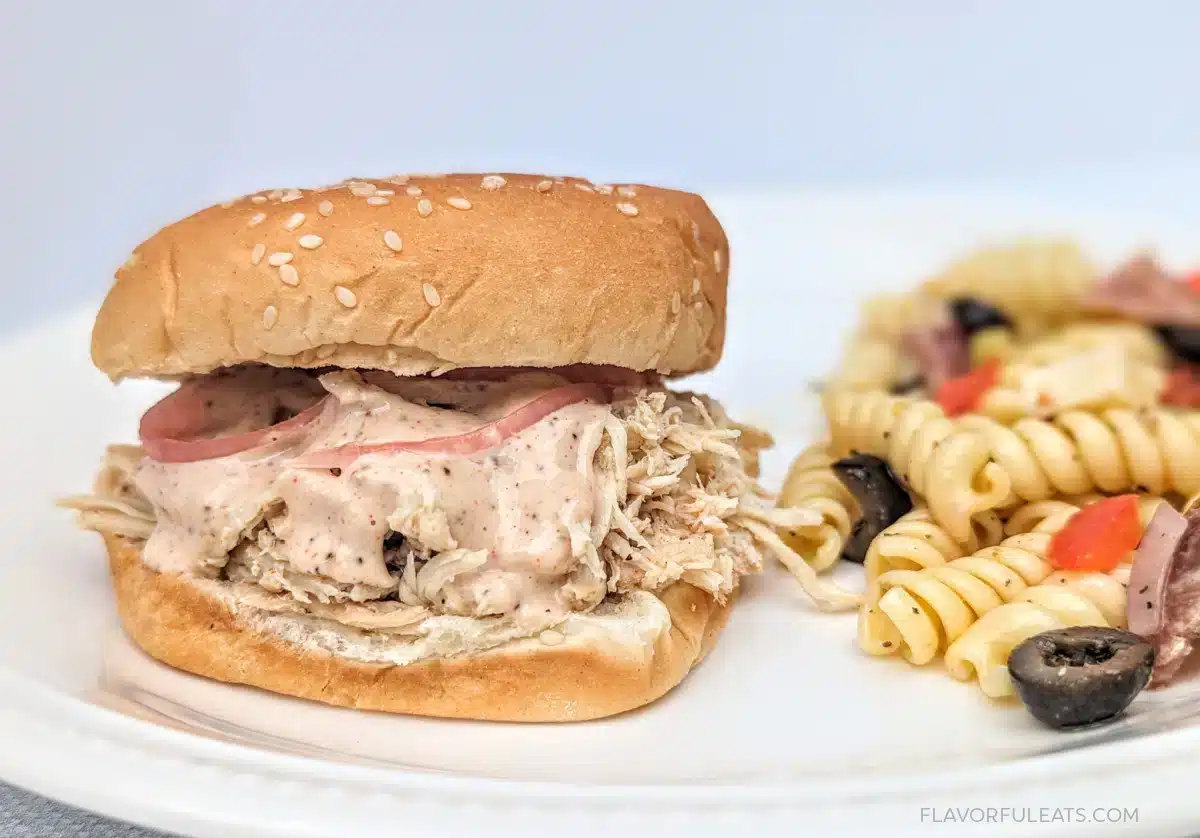 Shredded chicken sandwich with creamy sauce and pickles, served with pasta salad on a white plate.