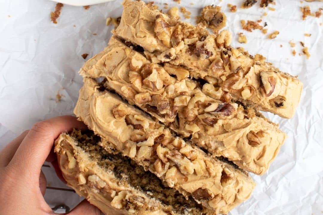 A hand holding slices of cake with frosting and chopped walnuts on top, on a white crumpled surface.