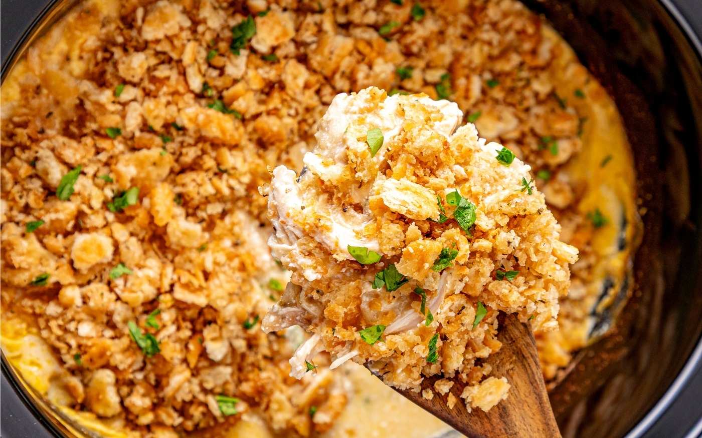 A wooden spoon scoops cheesy casserole topped with golden breadcrumbs and herbs from a baking dish.