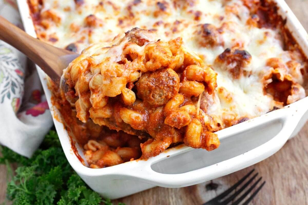 Baked pasta with meatballs and melted cheese being served from a white casserole dish with a wooden spoon.