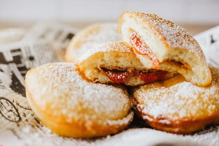 Three powdered sugar donuts, one cut open to reveal a red jam filling, rest on printed parchment paper.