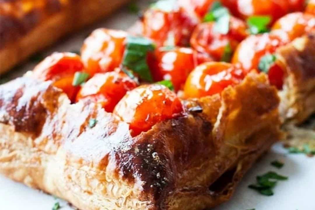 Rectangular puff pastry topped with roasted cherry tomatoes and fresh chopped herbs on a white surface.