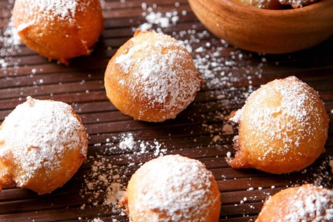 Round beignets dusted with powdered sugar on a wooden surface, with a wooden bowl in the background.