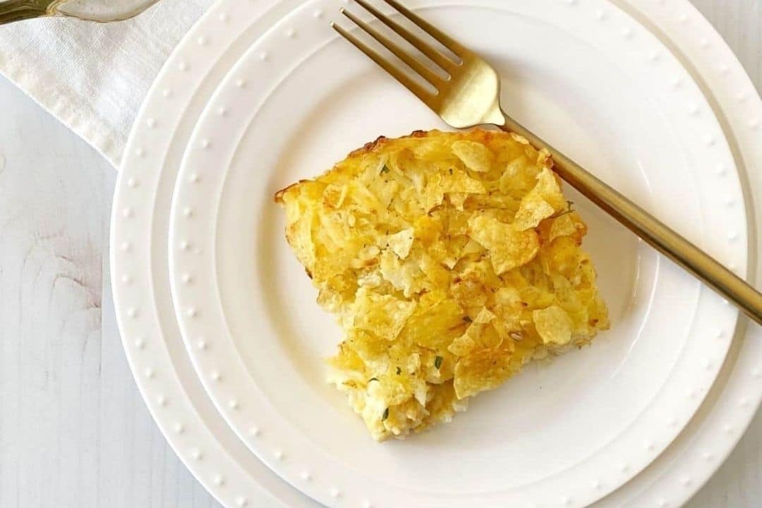 A serving of casserole topped with crushed potato chips on a white plate with a gold fork.