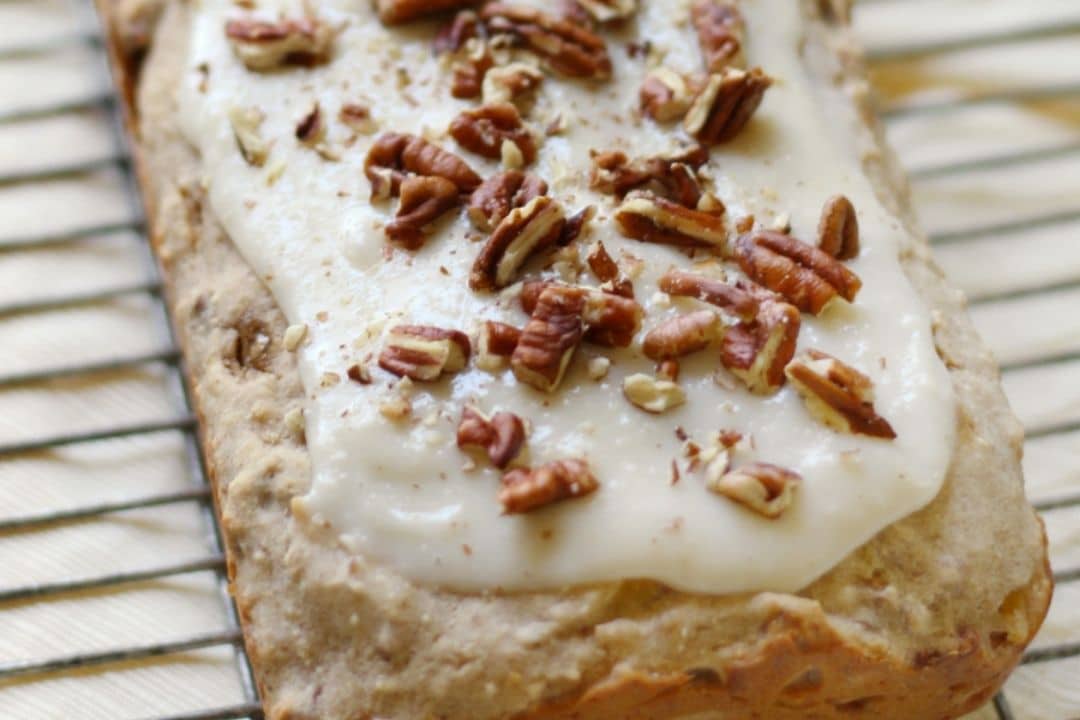 Loaf cake topped with white icing and chopped pecans, cooling on a wire rack.