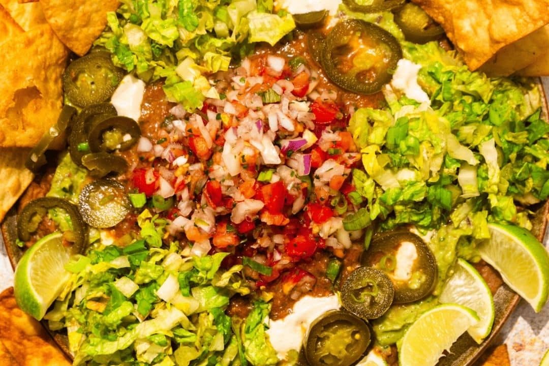 Colorful nachos topped with guacamole, jalapeños, lettuce, pico de gallo, sour cream, and lime wedges.