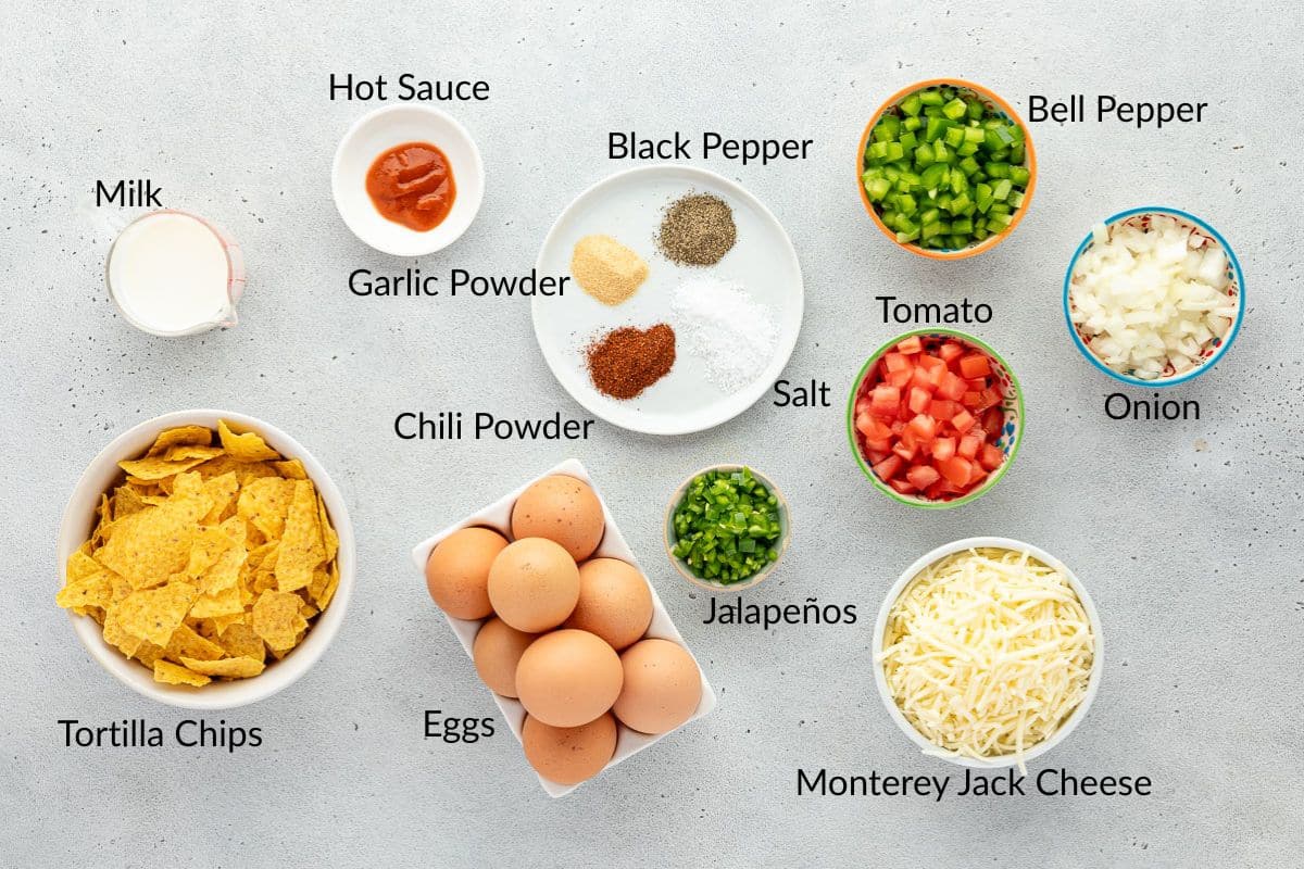 Ingredients for a recipe arranged on a counter, including eggs, tortilla chips, cheese, vegetables, and spices.