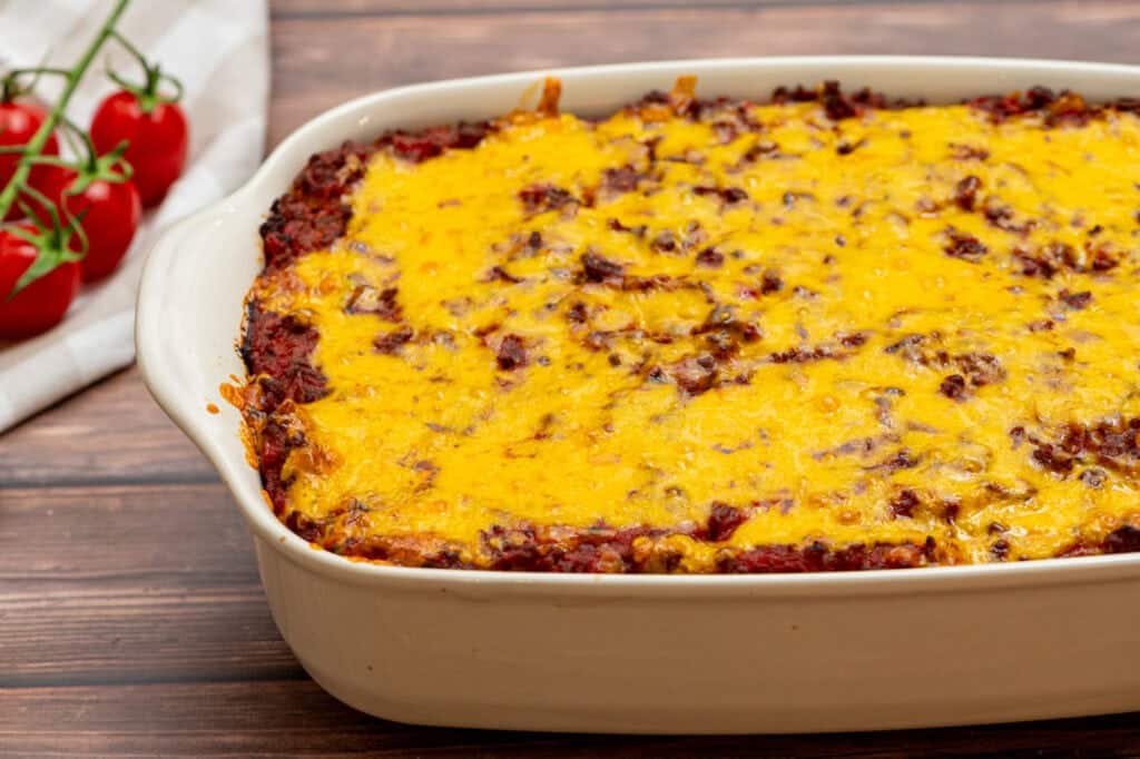 Cheesy casserole with a browned top in a white dish, with tomatoes on the vine nearby on a wooden table.