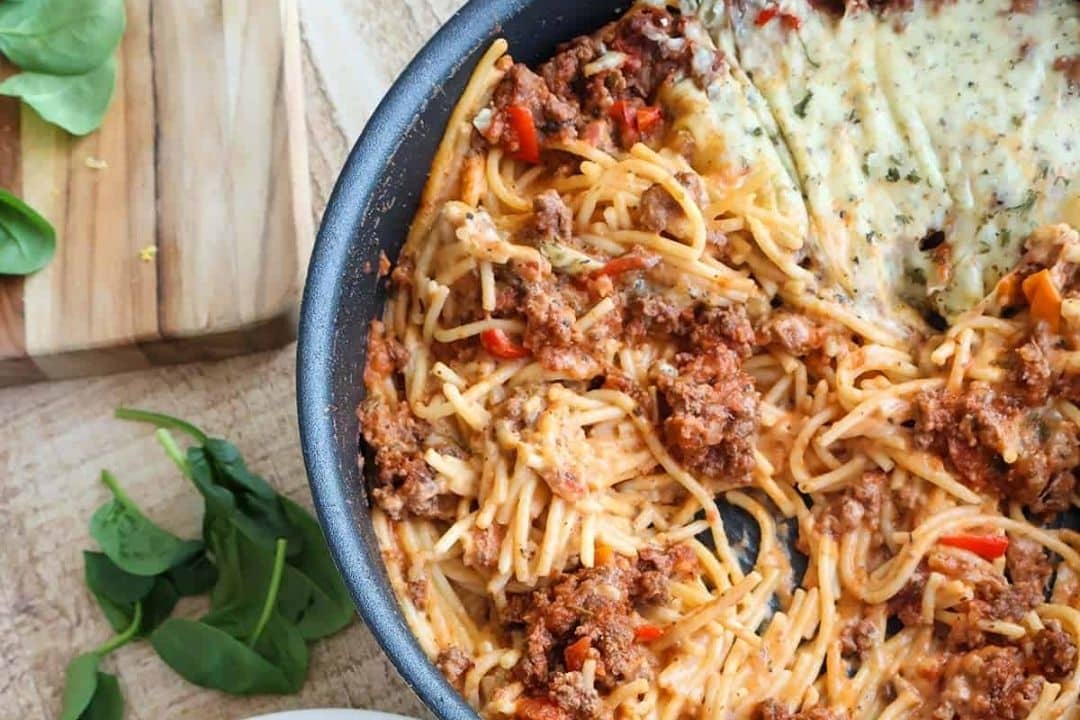 Baked spaghetti with ground meat, cheese, and tomato sauce in a skillet on a wooden surface.
