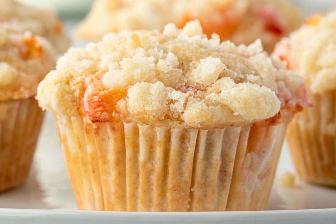 Close-up of a peach muffin with crumbly streusel topping on a white plate.