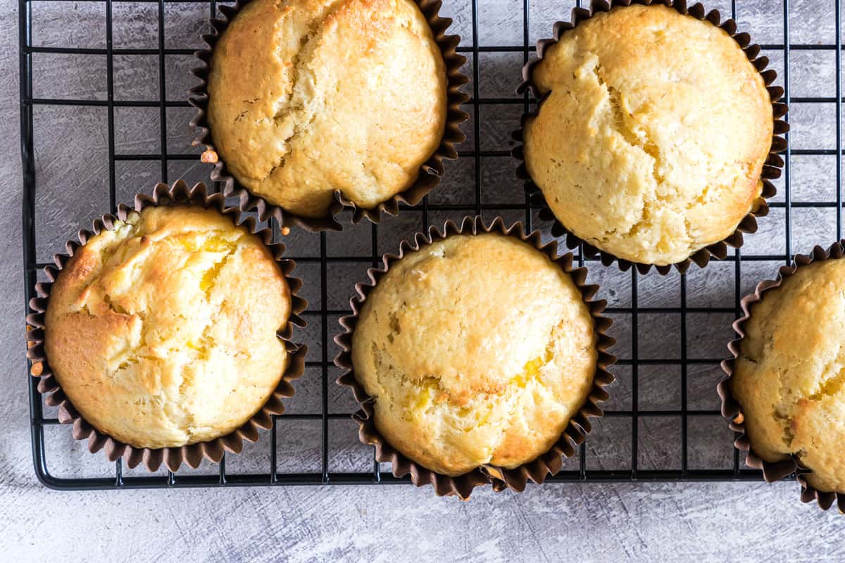 Five golden muffins in paper liners cooling on a black wire rack over a gray textured surface.