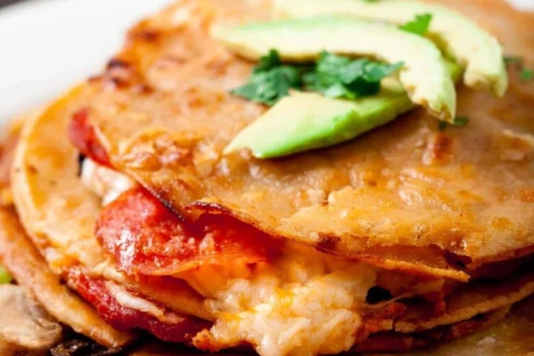 Cheesy quesadilla with tomato and avocado slices on top, garnished with cilantro on a plate.