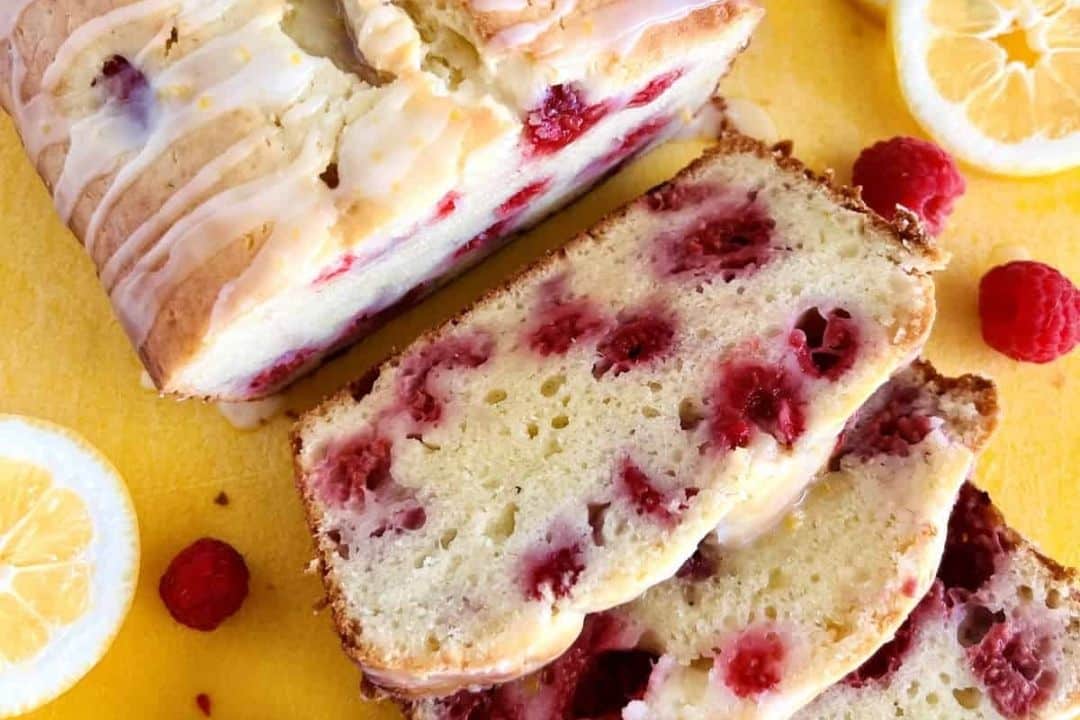 Sliced lemon raspberry loaf cake with icing, surrounded by fresh raspberries and lemon slices on a yellow surface.