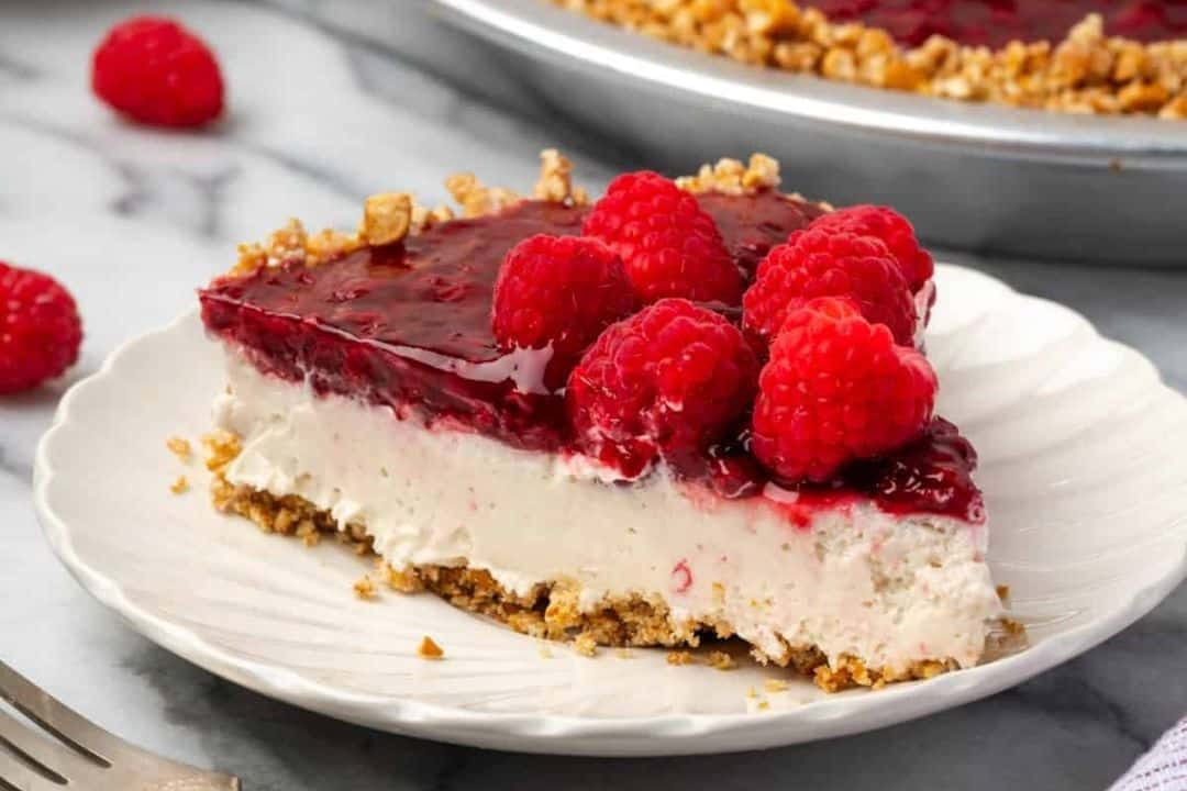 A slice of cheesecake with raspberry topping and fresh raspberries on a white plate, with a crumb crust.