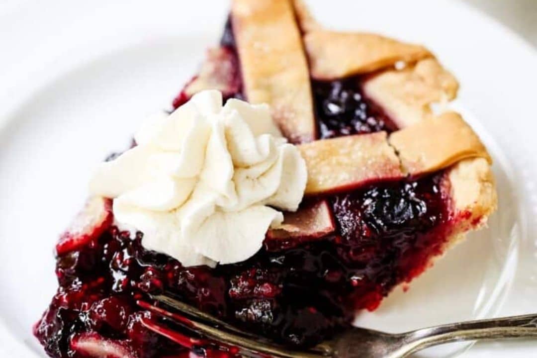 A slice of berry pie with lattice crust and whipped cream on a white plate with a fork.