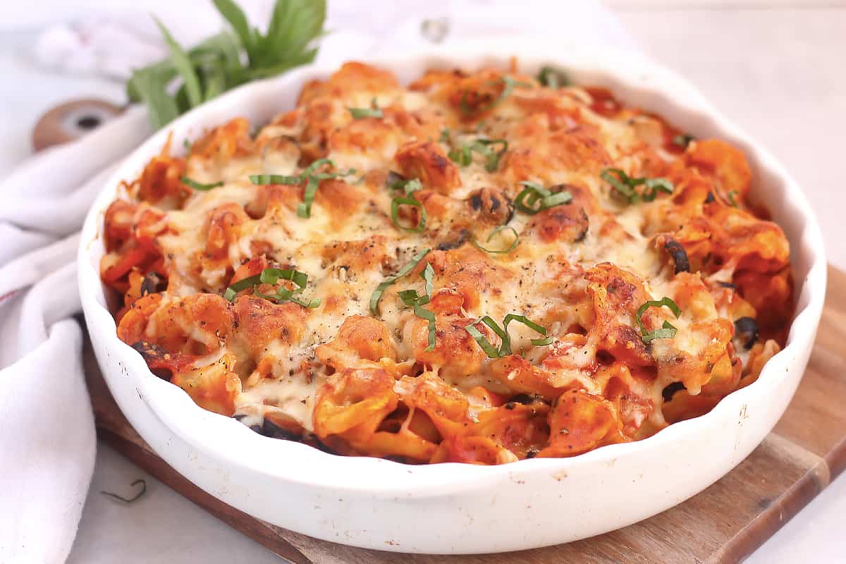 Baked pasta with melted cheese and fresh basil in a white dish on a wooden board.