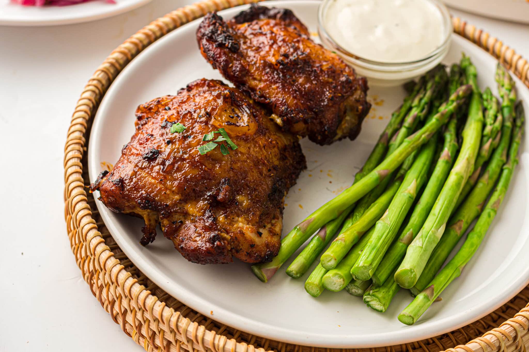 Grilled chicken thighs, asparagus, and a bowl of white dipping sauce on a round white plate.